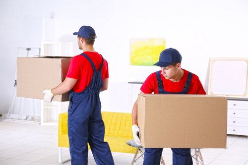 Two removal service workers dressed in red shirts, blue overalls, and blue caps are handling cardboard boxes during a house move inside a bright, modern home. The worker on the left, seen from behind, is holding a box with both hands and is standing near a yellow sofa with a textured fabric finish. The second worker on the right, who is facing slightly to the side, is kneeling or bent over as he lifts a large cardboard box, wearing white gloves for handling. The background features a white wall with framed abstract artwork, a white chest of drawers, and some decorative objects. The environment is well-lit with natural light, highlighting the clean and organized interior, typical of residential properties in Kingston upon Thames. This scene illustrates professional house removal services, including careful handling of household items such as boxes and small furniture, in a typical home setting within the London borough area, with subtle reference to local moving requirements.