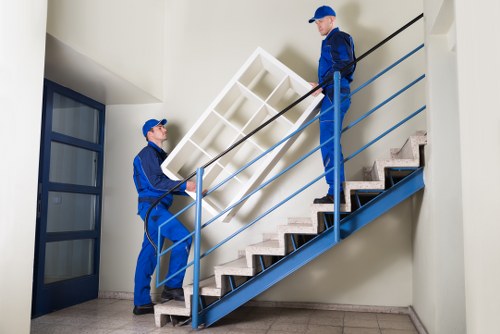 Two professional movers from Kingston upon Thames Man with Van are carrying a large, white wooden shelving unit up a staircase inside a residential building. The movers are dressed in matching blue uniforms and caps. One mover is positioned at the bottom of the stairs, lifting the shelving unit, while the other is higher up, guiding it onto the staircase railing. The staircase features dark wooden steps with a light grey tiled floor at the base and a modern blue metal handrail on the right side. To the left, there is a blue-framed glass door leading to another room, and the interior walls are painted white, providing a clean, neutral backdrop. Natural light illuminates the scene, highlighting the careful handling of the furniture, indicative of careful packing and moving services offered in the Kingston upon Thames area. This image reflects a professional home removal process, emphasizing careful furniture transportation within a typical residential property during a house move in southwest London.