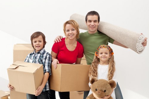 A family of four, consisting of a mother, father, a young boy, and a young girl, standing in a bright, white interior space with a plain background. The mother is wearing a red top and has short, red hair, while the father is dressed in a green t-shirt and is carrying a large rolled-up carpet on his shoulder. The young boy, positioned on the left, wears a blue and white checkered shirt and is holding two stacked cardboard boxes. The girl, on the right, has long, blonde hair with a colorful headband, and she is holding a light brown teddy bear. The family members are smiling, appearing cheerful and ready for a household move. In the foreground, there are additional cardboard boxes of various sizes, some sealed, indicating packing for a house relocation. The scene reflects a typical house move in Kingston upon Thames, with family-oriented elements and household items prepared for transport, fitting within the context of professional removals services at KingstonuponThamesManWithVan.co.uk.