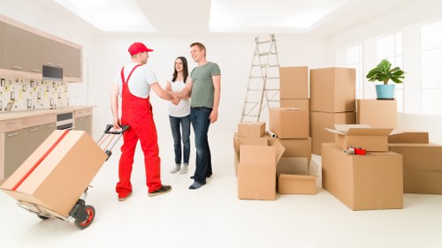 In a brightly lit, modern residential interior, a man in casual clothing is shaking hands with a house removal specialist dressed in a red uniform and cap, as another woman stands nearby, smiling and observing the interaction. The scene shows multiple large cardboard boxes, some open and others sealed, stacked along the right side of the room on the floor. A metal ladder is positioned against the wall, indicating ongoing packing or unpacking activities. To the left, part of a contemporary kitchen with wooden cabinetry and a tiled backsplash is visible. Natural light streams through large windows, highlighting the clean and organized space. The floor appears to be light wood or laminate, and a potted plant on top of a box adds a touch of greenery. This scene illustrates a professional house removal service, with attention to careful handling and packing of household belongings, characteristic of Kingston upon Thames area residential relocations conducted by Kingston upon Thames Man with Van.
