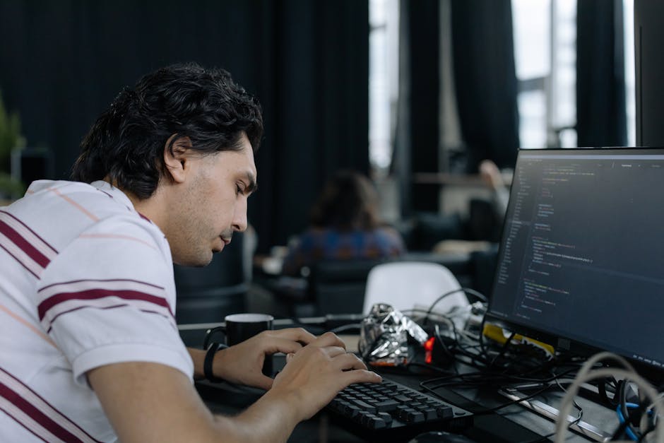 A person seen from behind working at a desk during a house removal process, with a laptop displaying lines of colorful code or technical data. The workspace includes two computer monitors, one of whic