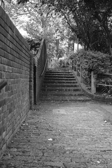 A black and white photograph showing a narrow outdoor stairway composed of brick steps ascending between two sides: on the left, a high brick wall, and on the right, a low brick wall topped with a met