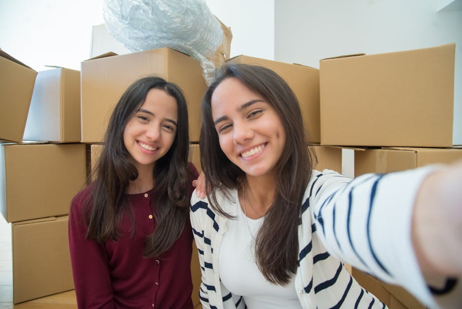 Two young women with long, dark hair are inside a property, surrounded by numerous packed cardboard boxes stacked on top of each other, likely prepared for a house removal. One woman is wearing a maro