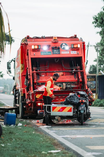 A worker wearing a high-visibility yellow vest and dark clothing is operating a waste disposal vehicle, positioned inside a driveway or street area at dusk or night. The vehicle is equipped with vario